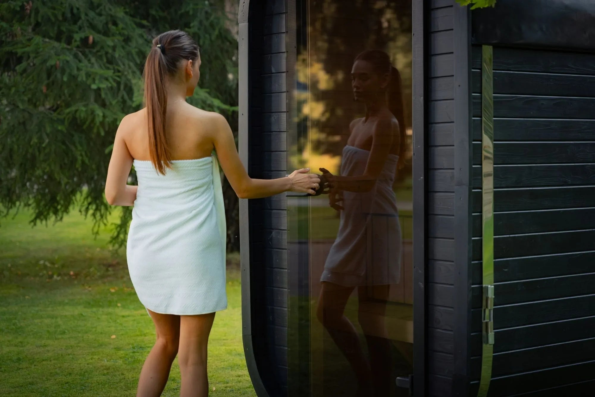 Frau im Handtuch betritt eine schwarze Design-Sauna mit Glasfront im Garten – architektonische Wellnesslösung im Einklang mit Natur und Umgebung.