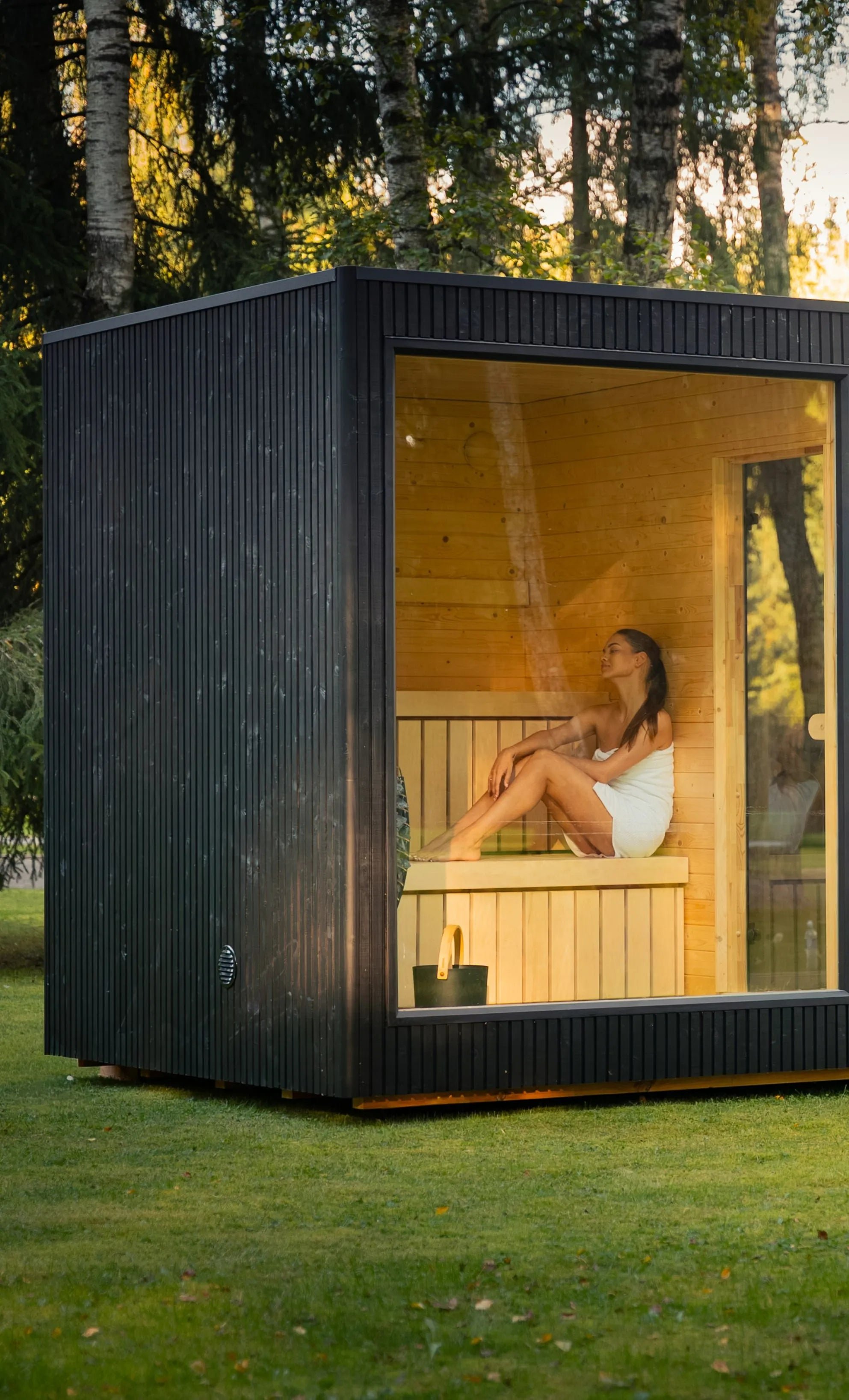 Schwarze Design Gartensauna mit großer Glasfront und integriertem Hot Tub in einer grünen Parklandschaft.