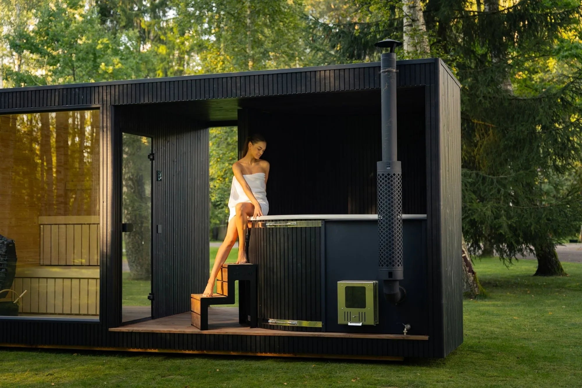 Frau im Handtuch betritt die Celcium Taurus Sauna aus Ebenholz und Glas im Wald und wechselt anschließend in den integrierten Hot Tub – luxuriöse Kombination aus Sauna und Whirlpool in natürlicher Umgebung.